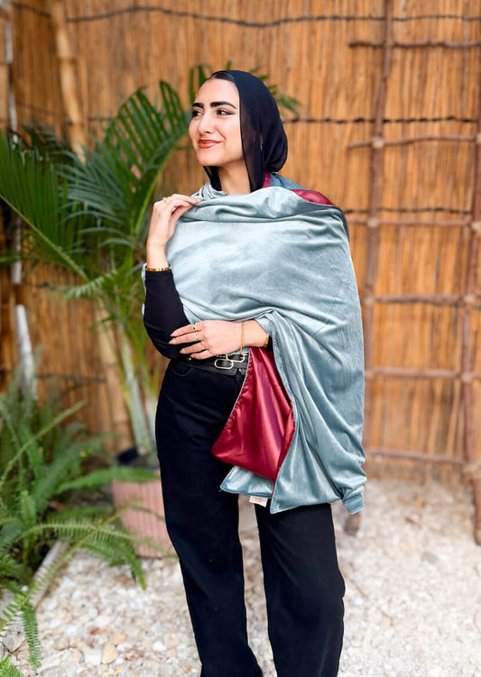 Woman holding a light blue shawl against a natural background with plants and a bamboo wall.