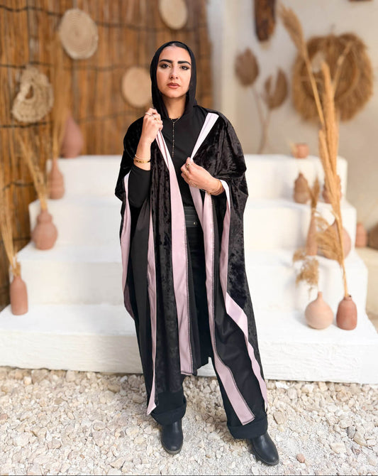Woman in a black and pink abaya standing in a decorative outdoor setting with plants and stones.