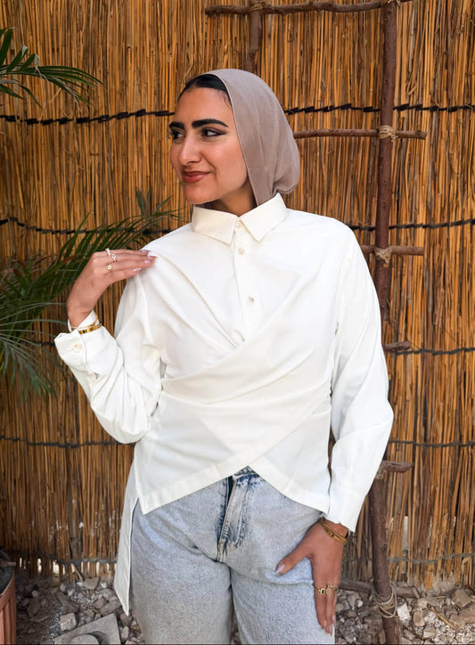 Woman wearing a white shirt and light gray hijab standing against a thatched wall.