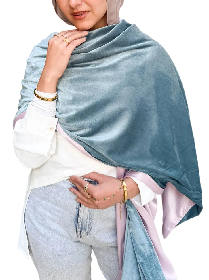 Woman wearing a light blue shawl over her shoulders with a wooden wall and plant in the background